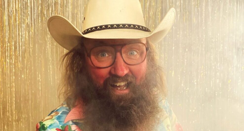 Onkel Reje med hvid cowboyhat, briller og langt gråt fuldskæg foran et gyldent glittergardin